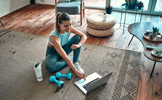 A sporty woman in sportswear is sitting on the floor with dumbbells and a protein shake or a bottle of water and is using a laptop at home in the living room. Sport and recreation concept.